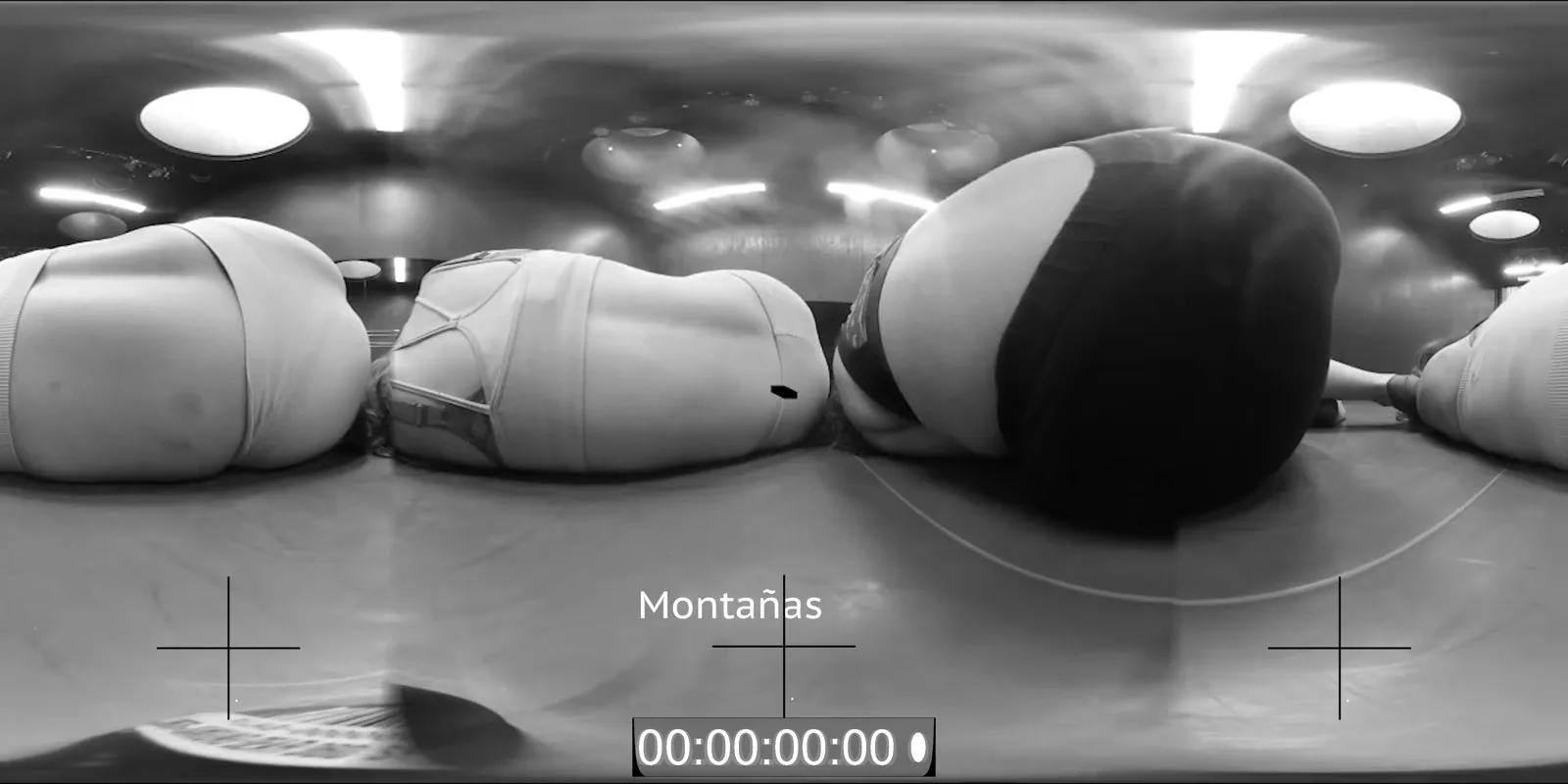 Fotogramma 360 in bianco e nero di una sala prove: tre corpi distesi in primo piano, crocini di registro e la scritta Montañas con il suo timecode