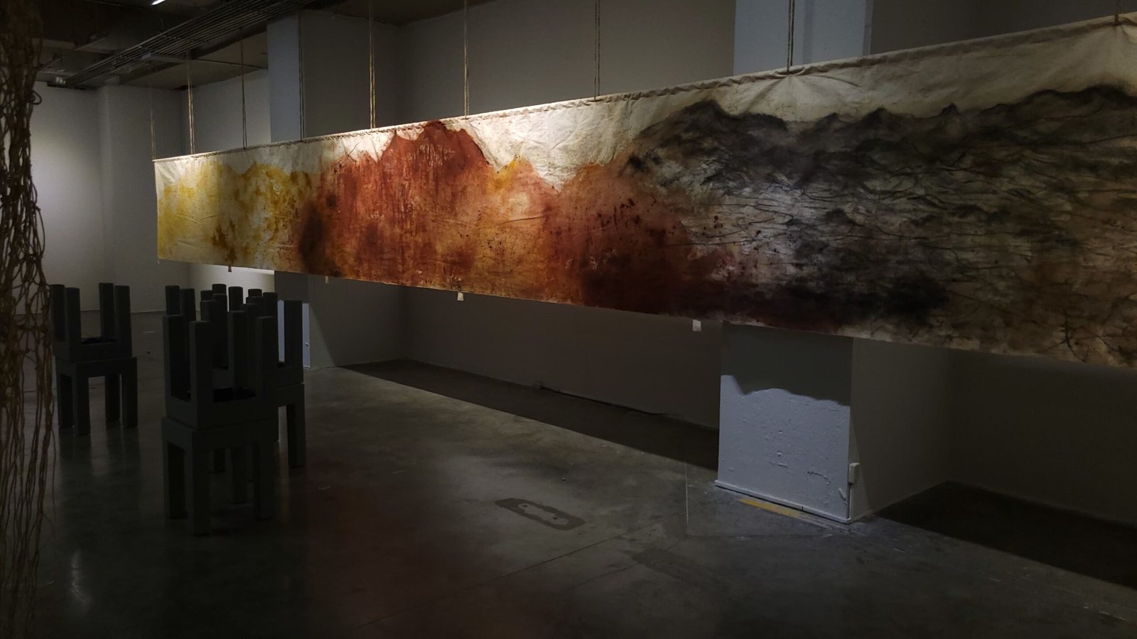 Tela larga colgada de cuerdas en una sala oscura, teñida con manchas amarillas, rojas y negras que dibujan una línea de montañas; debajo, taburetes de madera oscura.