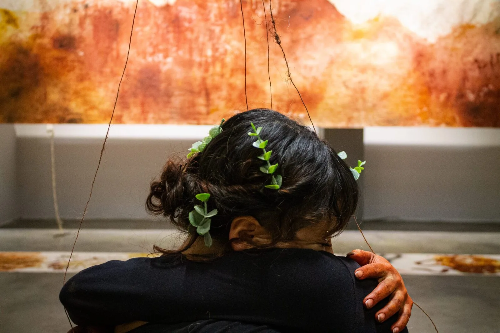 Due persone abbracciate, di spalle; una ha foglie verdi tra i capelli raccolti. Sullo sfondo, il telo tinto di arancione e ocra.