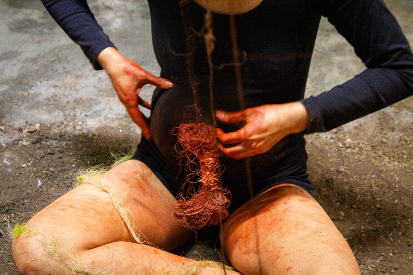 Manos manchadas de pigmento rojo sostienen un mechón de fibra teñida que cuelga de un hilo, sobre un cuerpo sentado en el suelo de tierra.