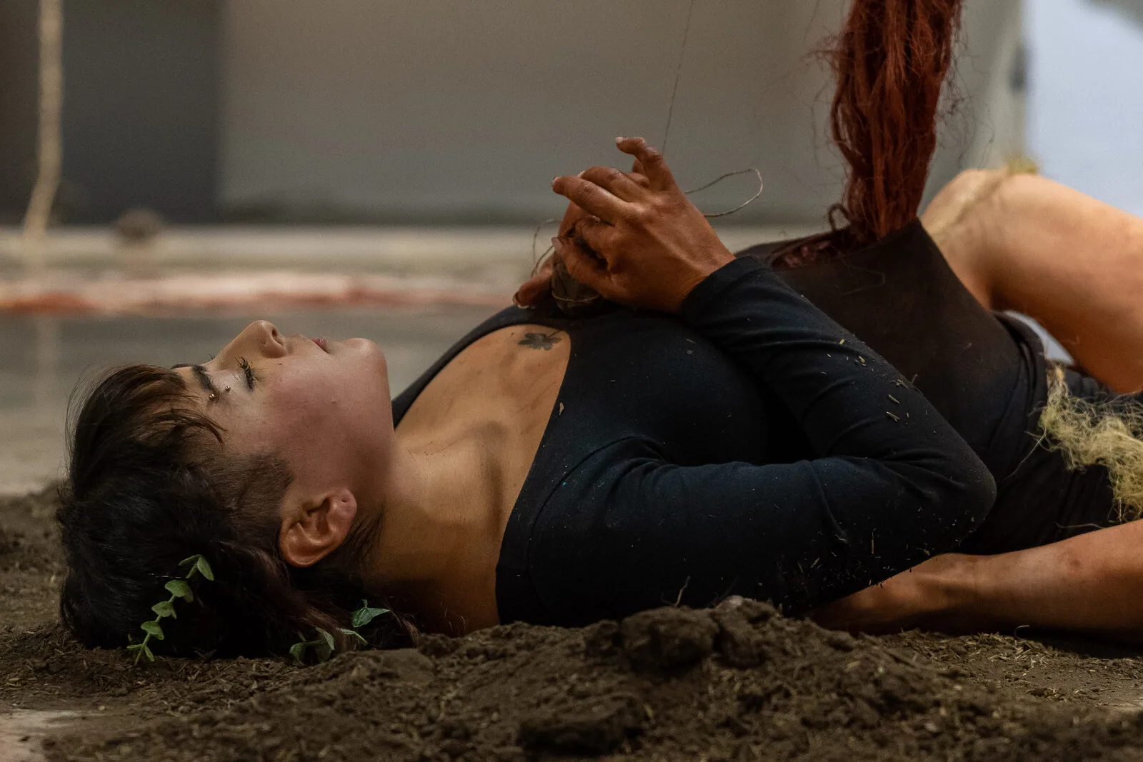 Una performer distesa a pancia in su sulla terra smossa, con foglie verdi tra i capelli e le mani incrociate sul petto.