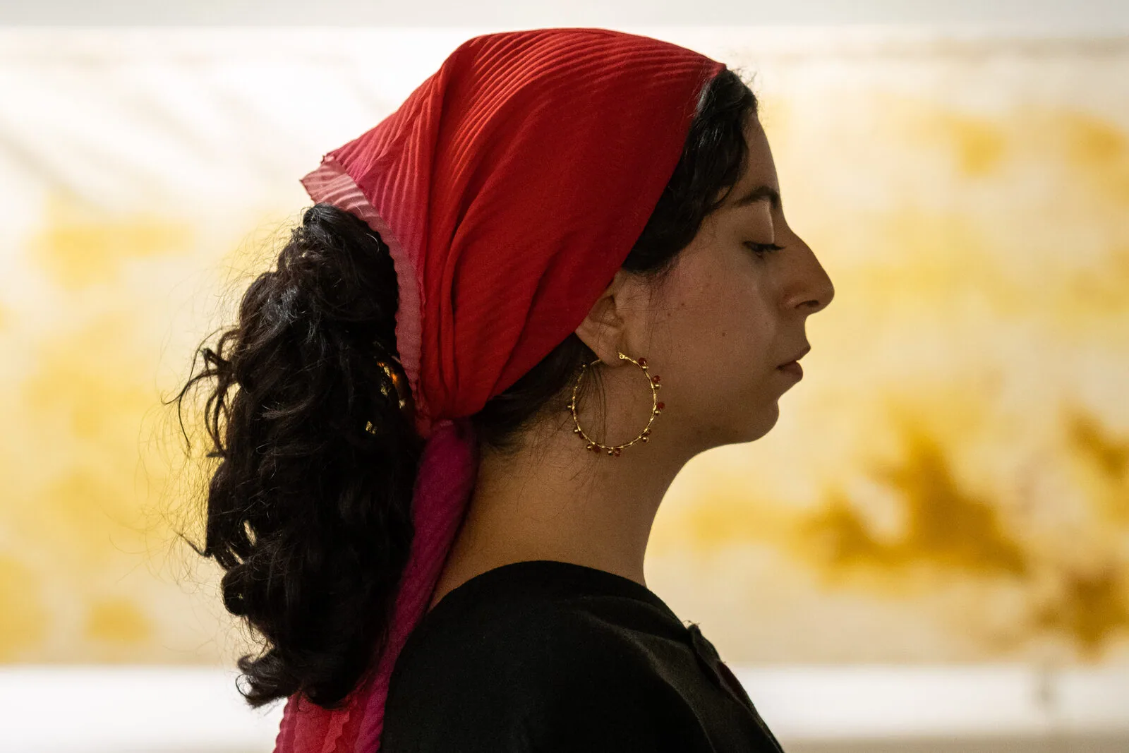 Profilo di una performer con un fazzoletto rosso in testa e orecchini a cerchio dorati, gli occhi chiusi, davanti al telo tinto di giallo.