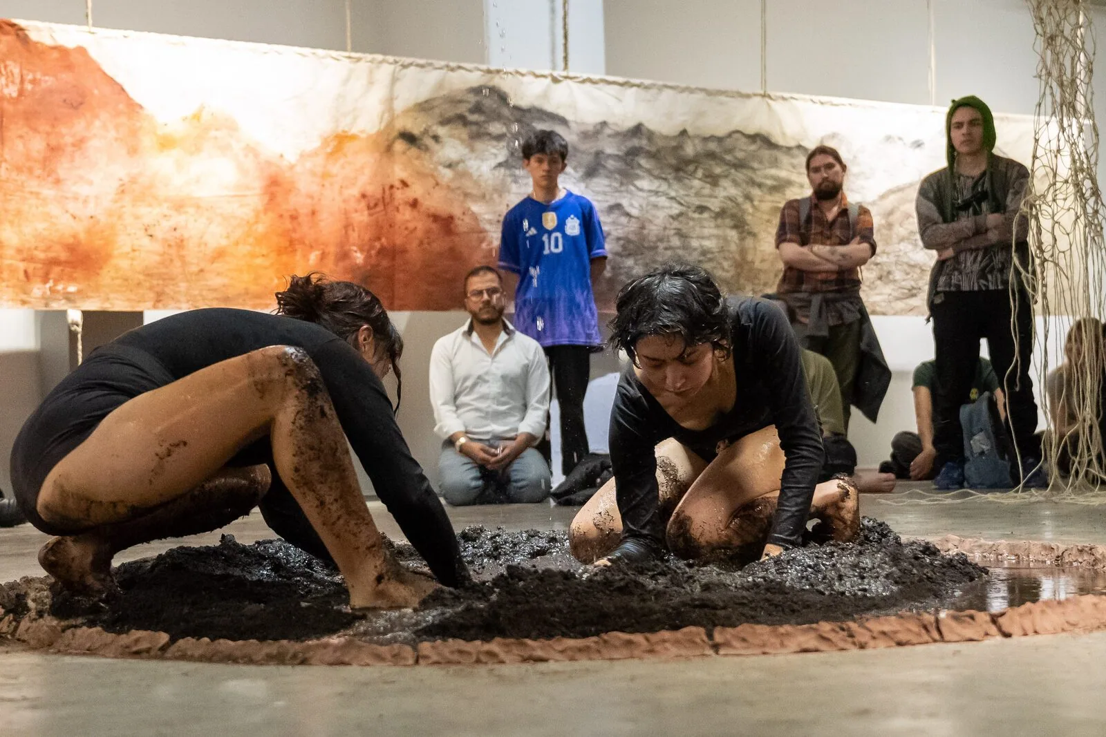 Due performer scavano con le mani nel fango dentro il cerchio di terra. Il pubblico guarda seduto a terra e in piedi; dietro, il telo tinto di arancione, nero e ocra.