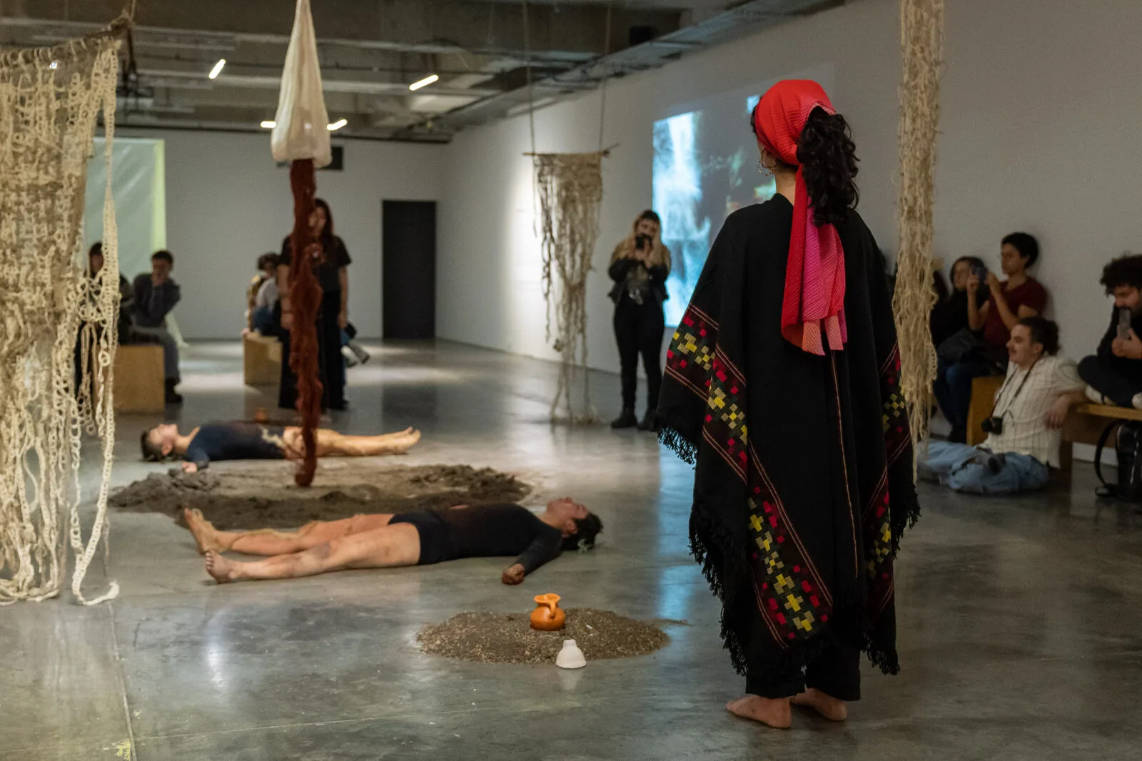 Campo lungo della sala: una persona con una ruana nera e un fazzoletto rosso osserva due performer distesi a terra tra cerchi di terra. Dal soffitto pendono tessuti di fibra e il pubblico circonda la scena.