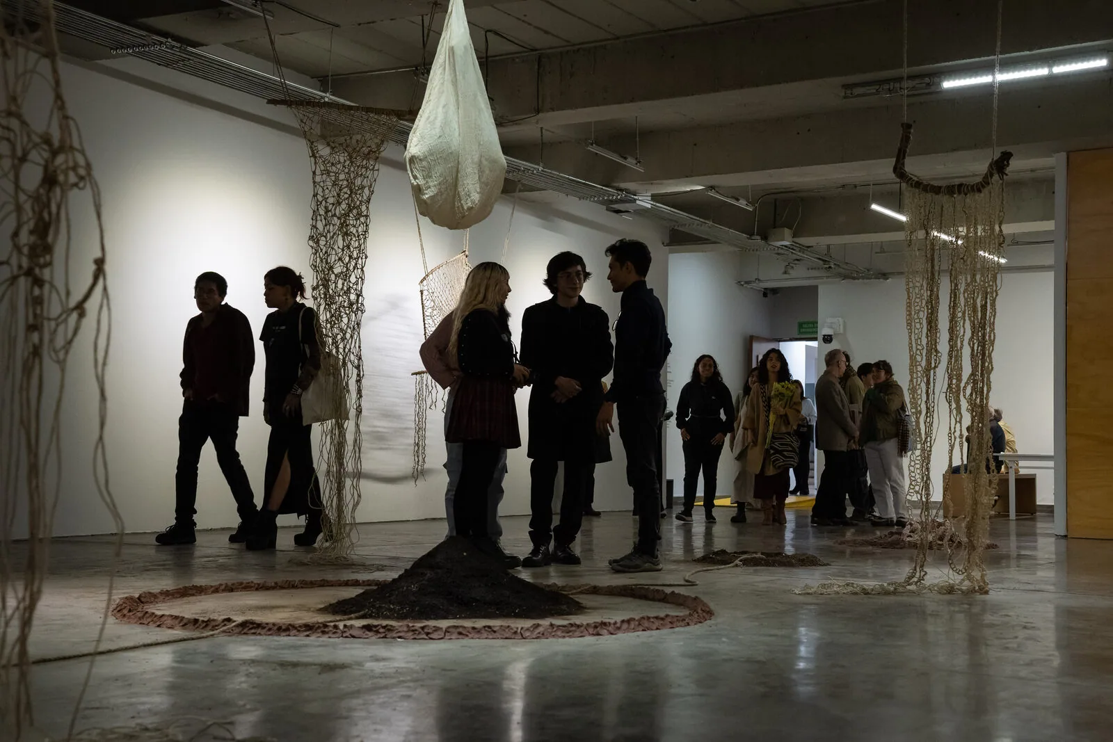 La sala in penombra: dal soffitto pendono un bozzolo di stoffa bianca e diversi tessuti di fibra; a terra, un cumulo di terra nera circondato da un anello di fango, e visitatori che attraversano lo spazio.