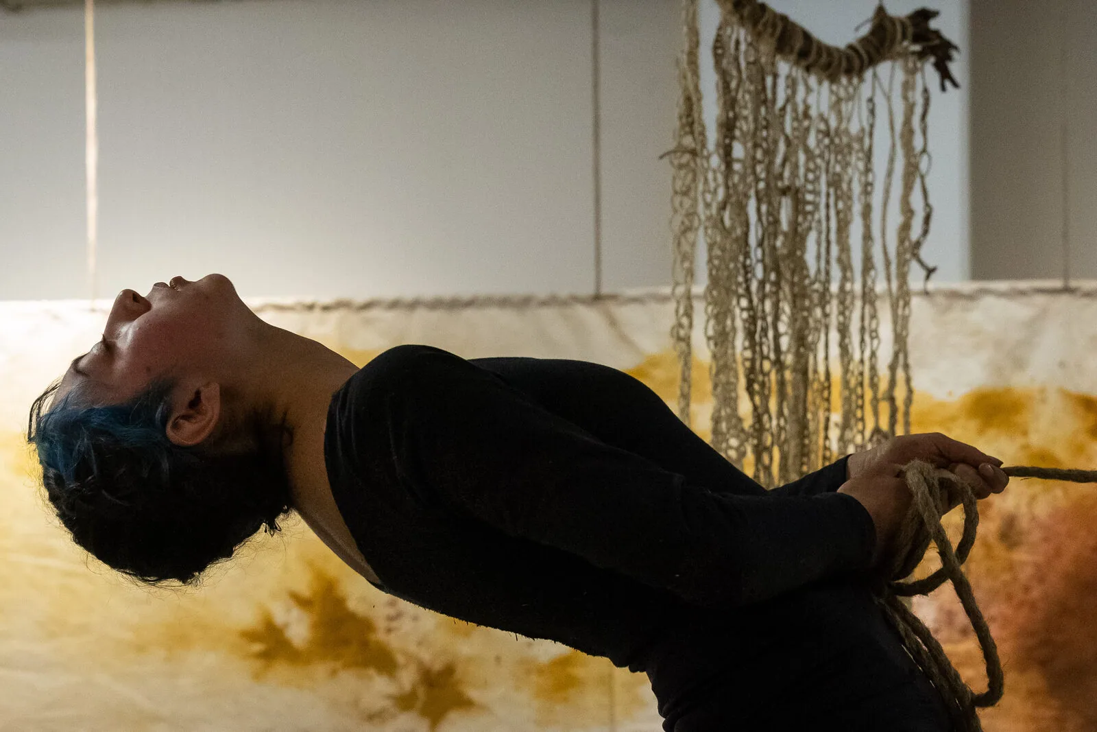 Una performer si inarca all'indietro tenendo una corda di fibra, davanti al telo tinto di giallo con la curcuma e ai cordoni intrecciati che scendono dal soffitto.