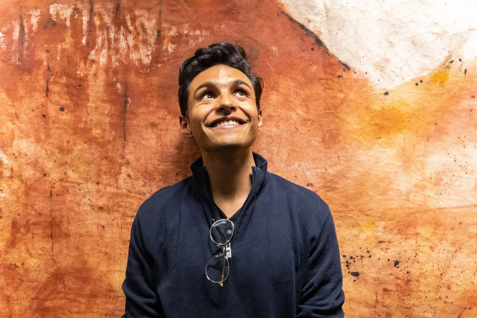 David Vega, con un maglione scuro e gli occhiali appesi al collo, guarda in alto sorridendo, appoggiato al telo tinto di ocra e di rosso.