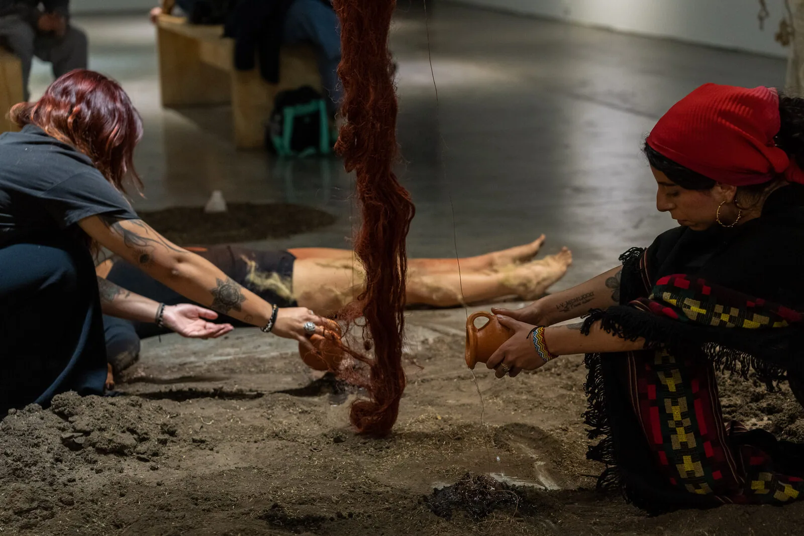 Due donne inginocchiate sulla terra: una versa acqua da un vaso di terracotta e l'altra la raccoglie con la mano aperta, accanto a una treccia di fibra rossa che scende dal soffitto e a un corpo disteso sullo sfondo.