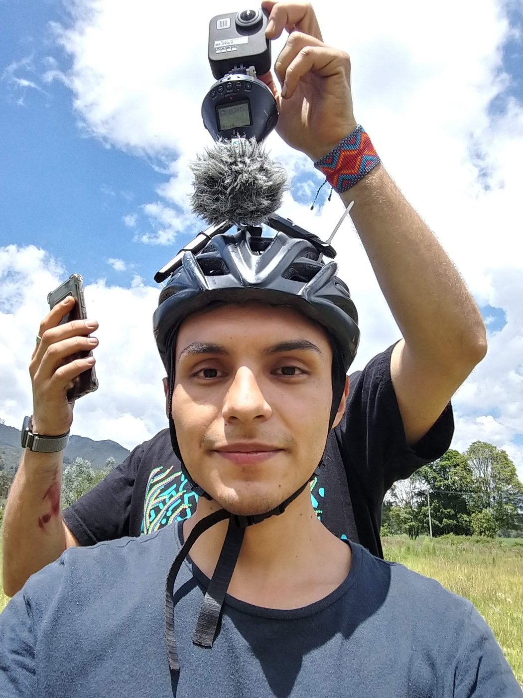 David con casco de ciclismo y, montados encima, una cámara 360, una grabadora de audio y un micrófono con paravientos; en la otra mano sostiene un celular, en un campo abierto.