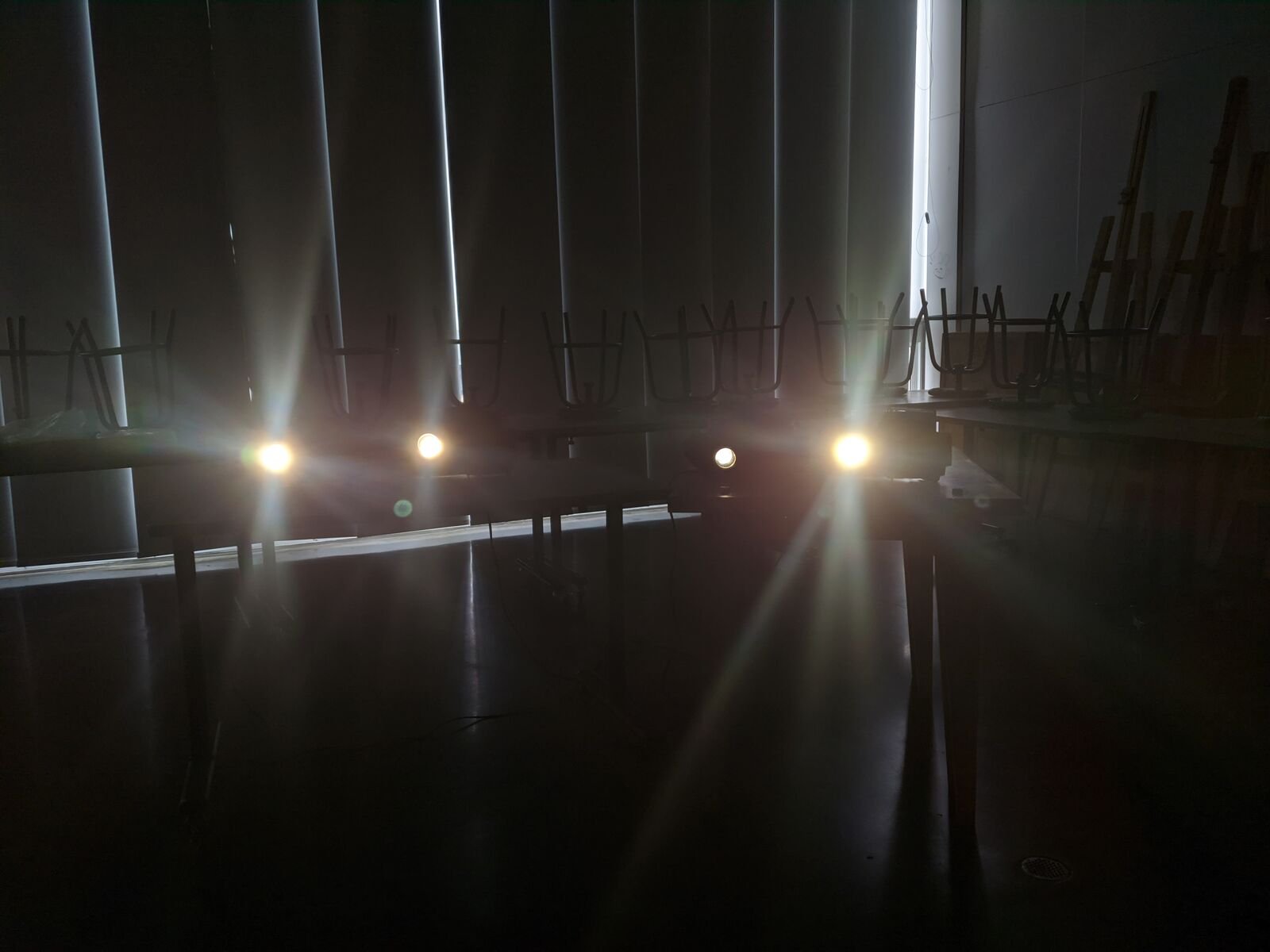 Cuatro proyectores en fila con las lámparas encendidas en un salón a oscuras, con persianas al fondo y sillas volteadas sobre las mesas.