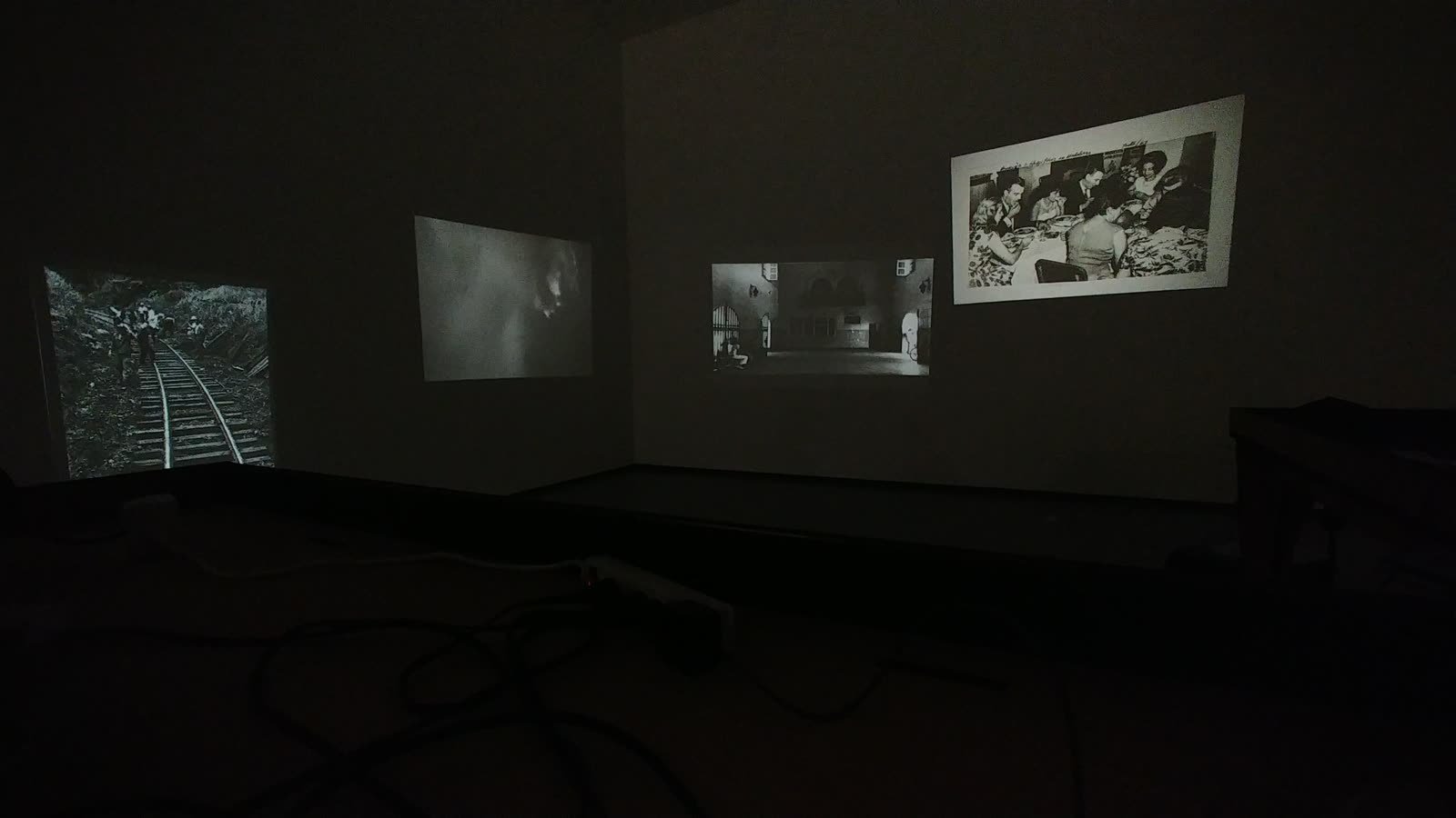 Sala oscura con cuatro proyecciones en blanco y negro sobre dos muros; en una se ven trabajadores junto a una vía férrea.